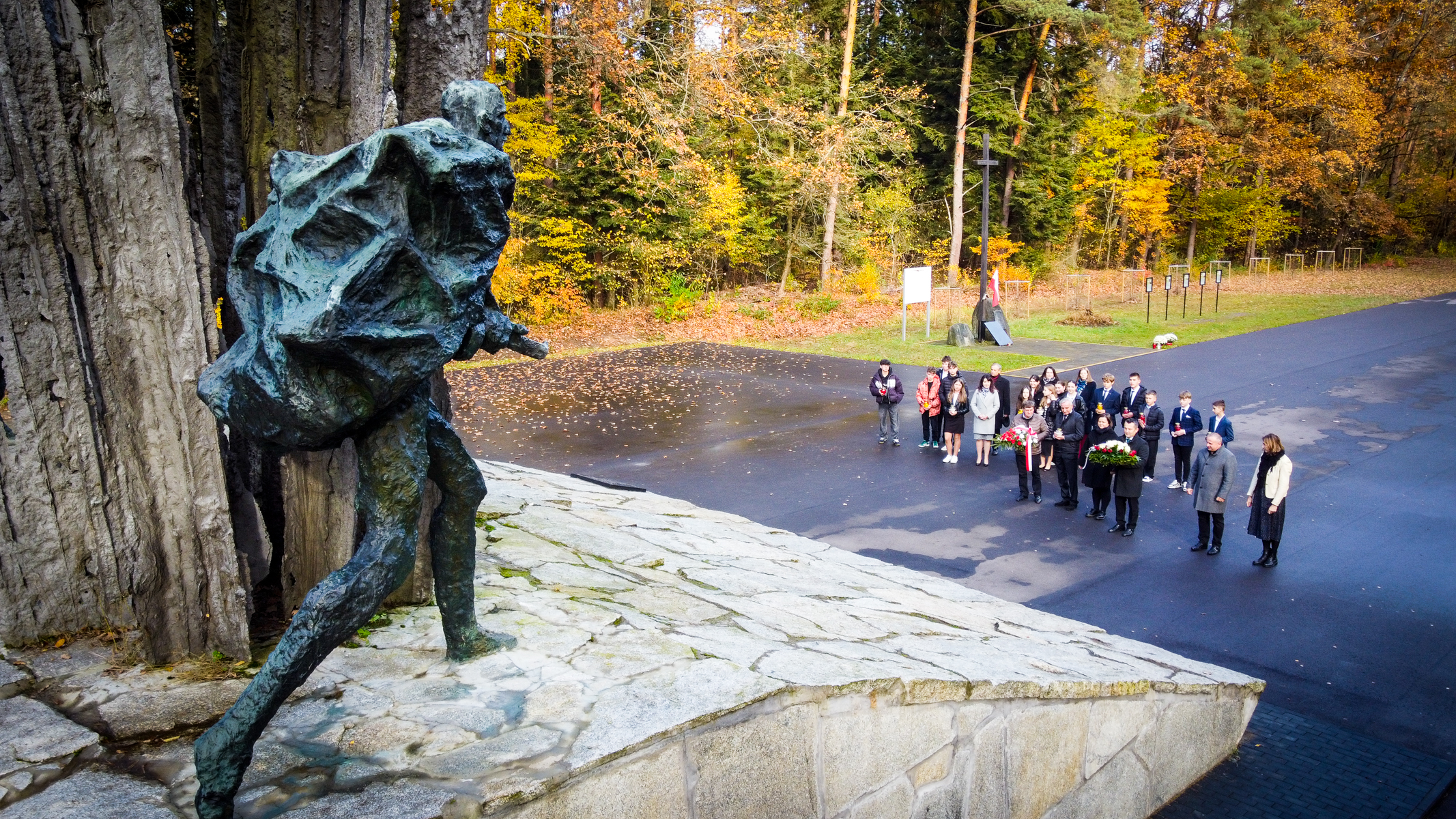 Rzeźba postaci z plecakiem na tle kamiennego podestu, grupa ludzi z kwiatami na asfaltowym placu, jesienne drzewa w tle.