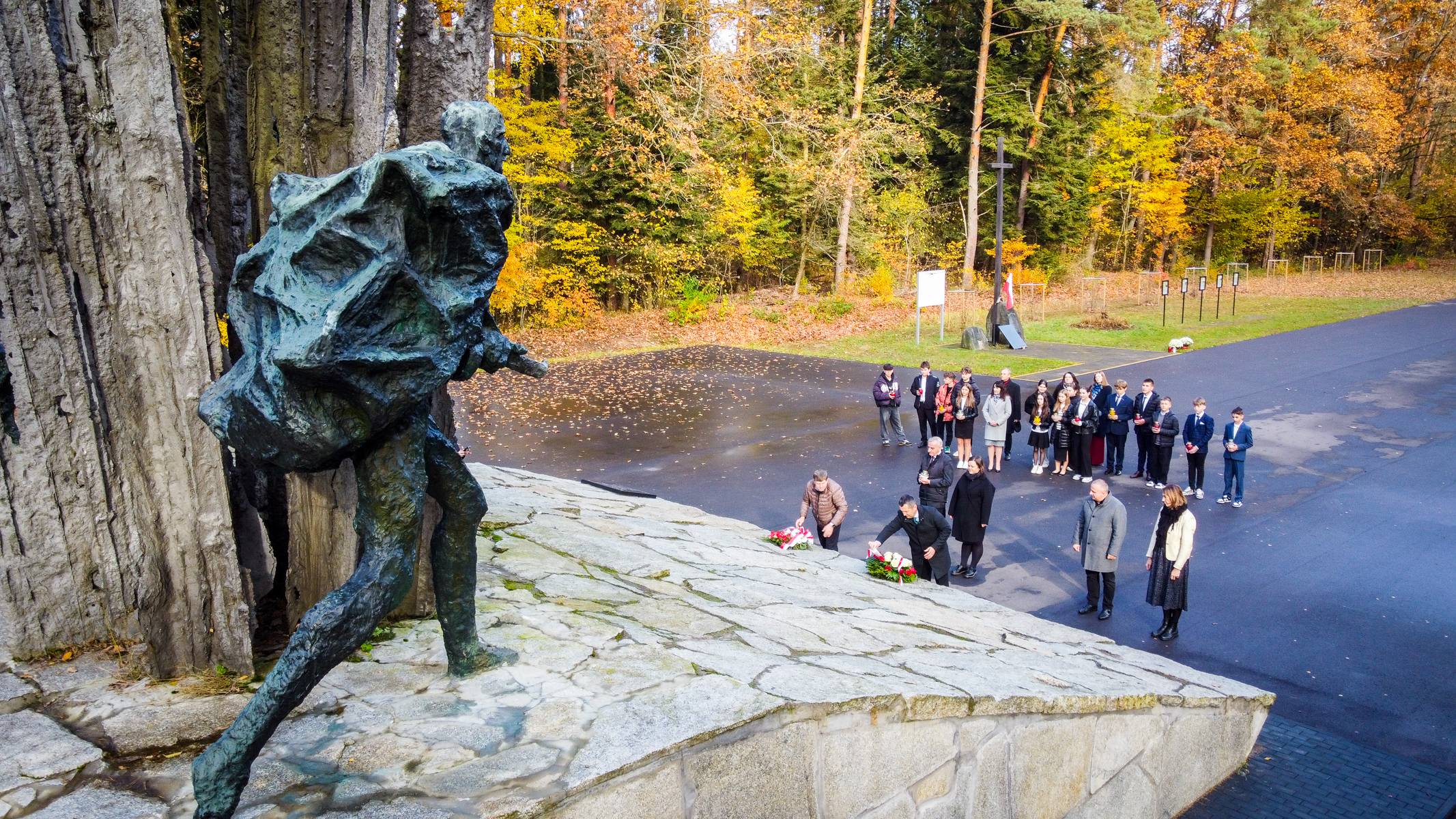 Brązowa rzeźba mężczyzny niosącego ciężki ładunek na plecach, grupa ludzi składająca kwiaty i znicze na kamiennym pomniku, jesienne drzewa w tle.