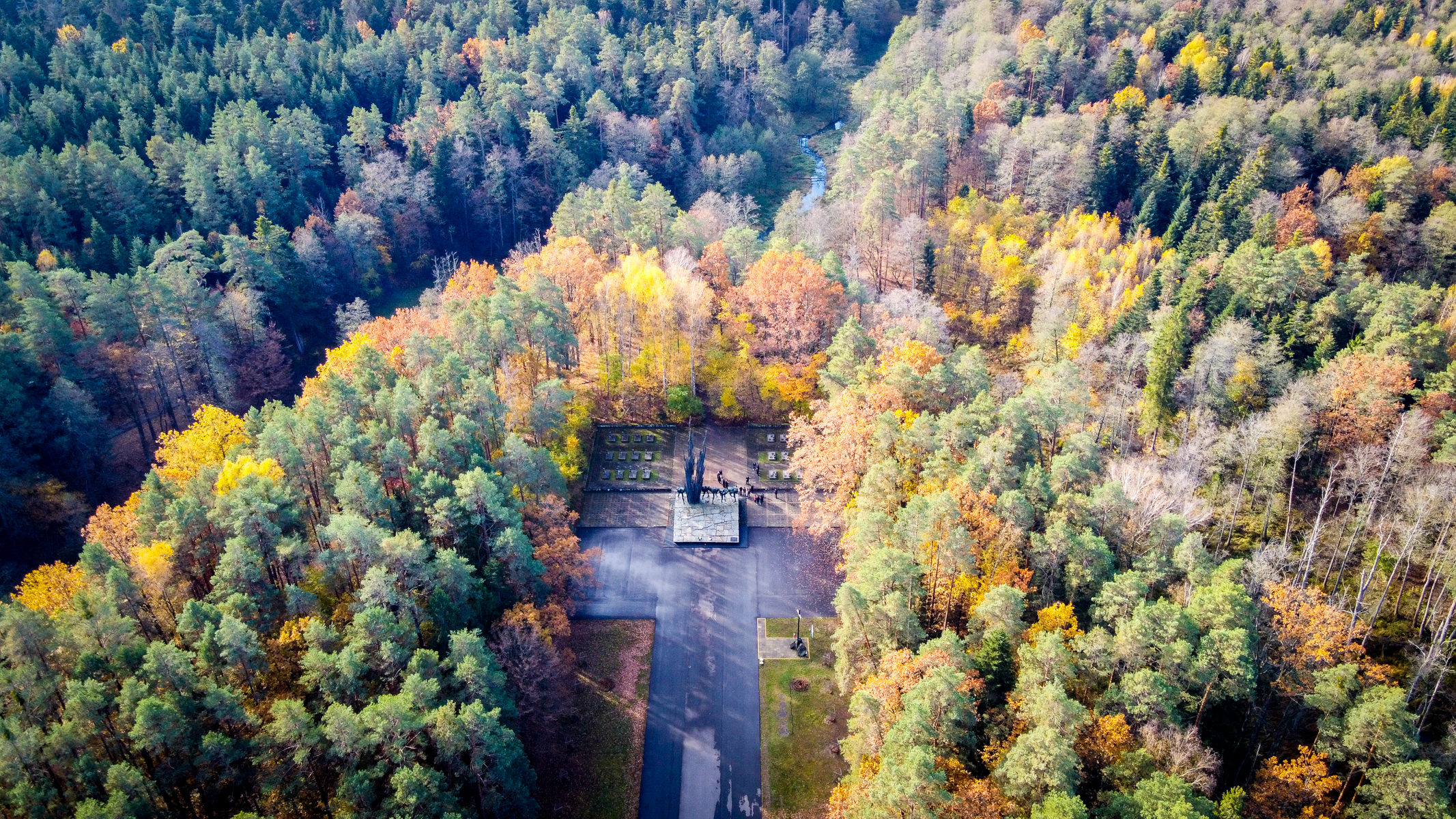 Pomnik na prostokątnej platformie otoczony kolorowym lasem z przewagą zieleni, żółci i pomarańczy, widok z lotu ptaka, asfaltowa droga prowadząca do pomnika.