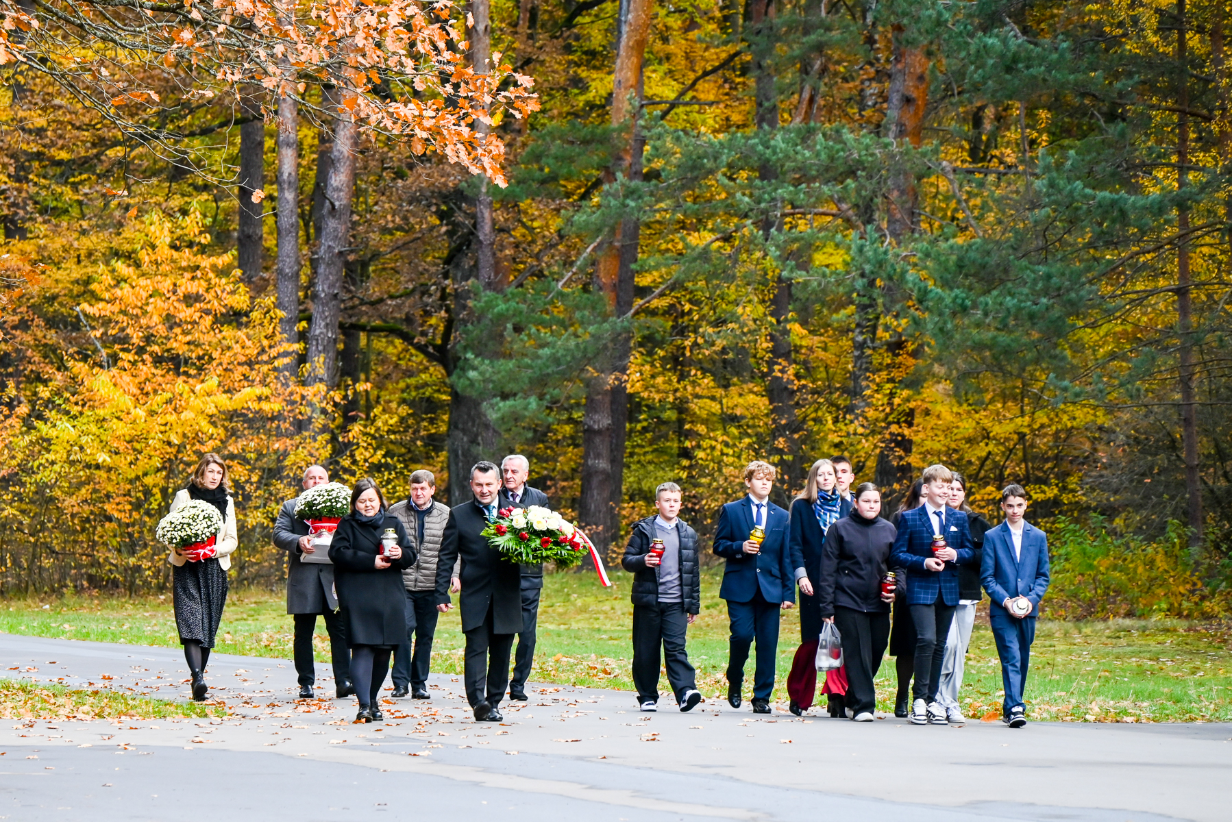 Grupa ludzi w jesiennym lesie, niektórzy niosą kwiaty i znicze, ubrani w ciemne i formalne stroje, wokół kolorowe liście i drzewa.