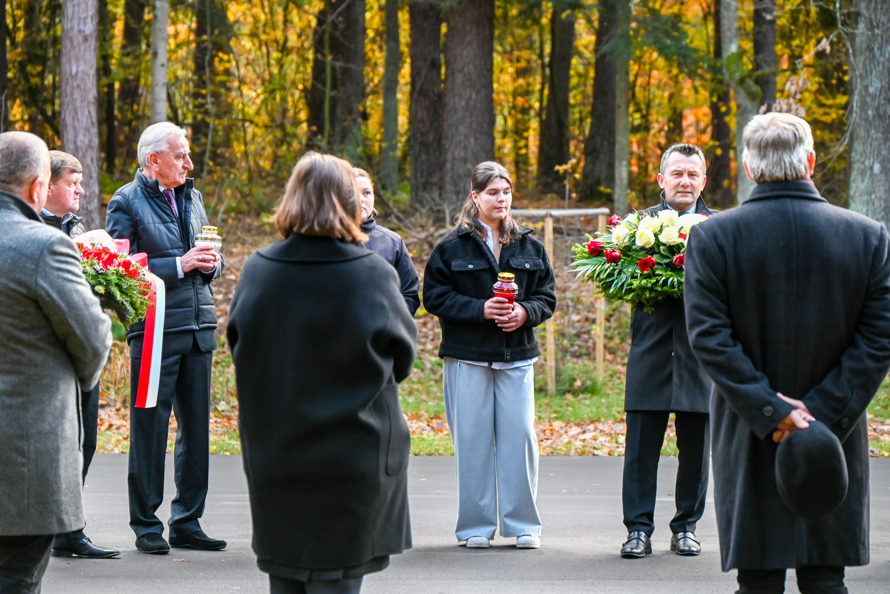 Grupa ludzi w jesiennym lesie, mężczyzna z wieńcem z białych i czerwonych róż, kobieta z czerwonym zniczem, inni w ciemnych płaszczach, liście na ziemi.