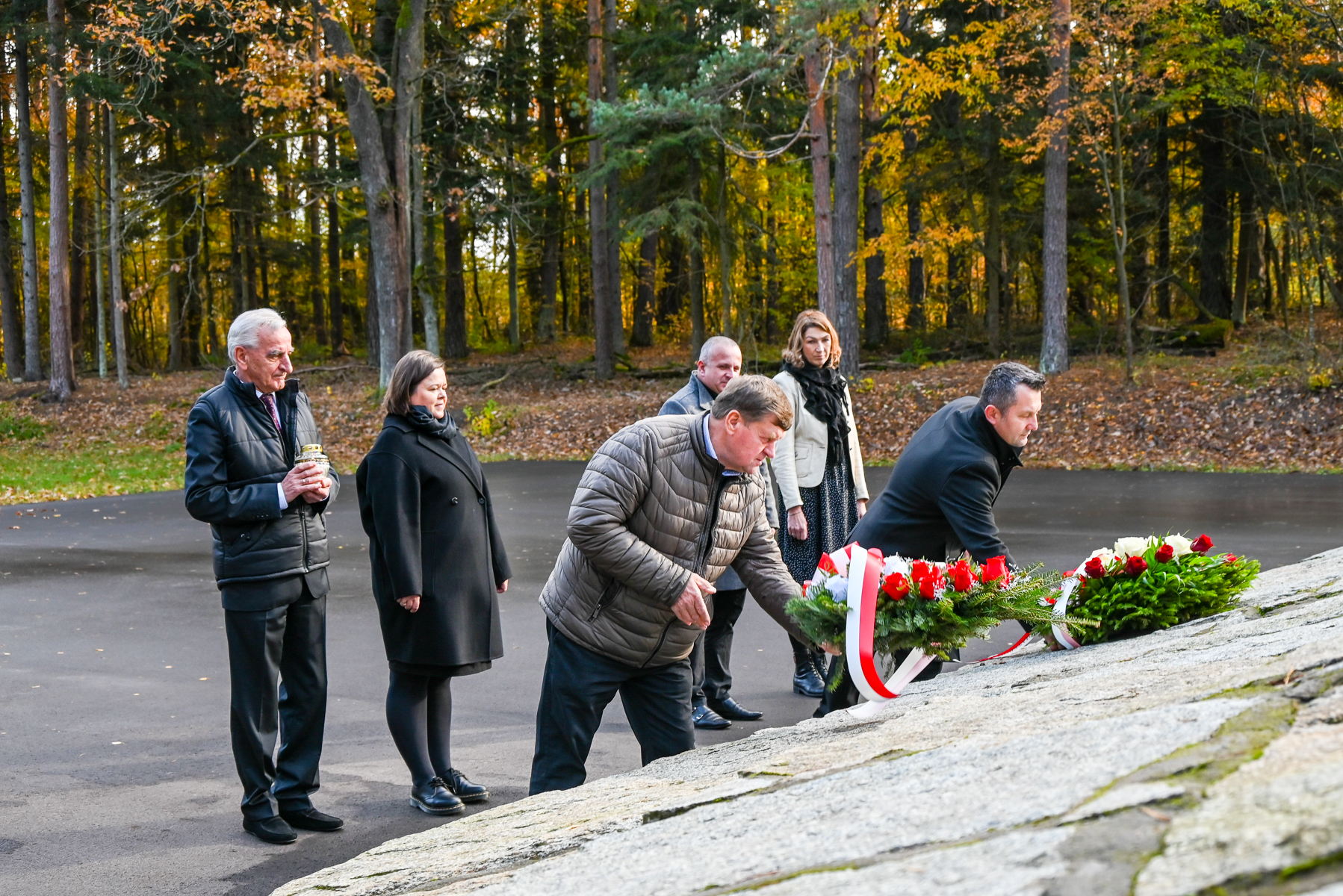 Pięć osób w jesiennym lesie układa wieńce z czerwonych i białych kwiatów na kamiennym pomniku, jedna osoba trzyma znicz.