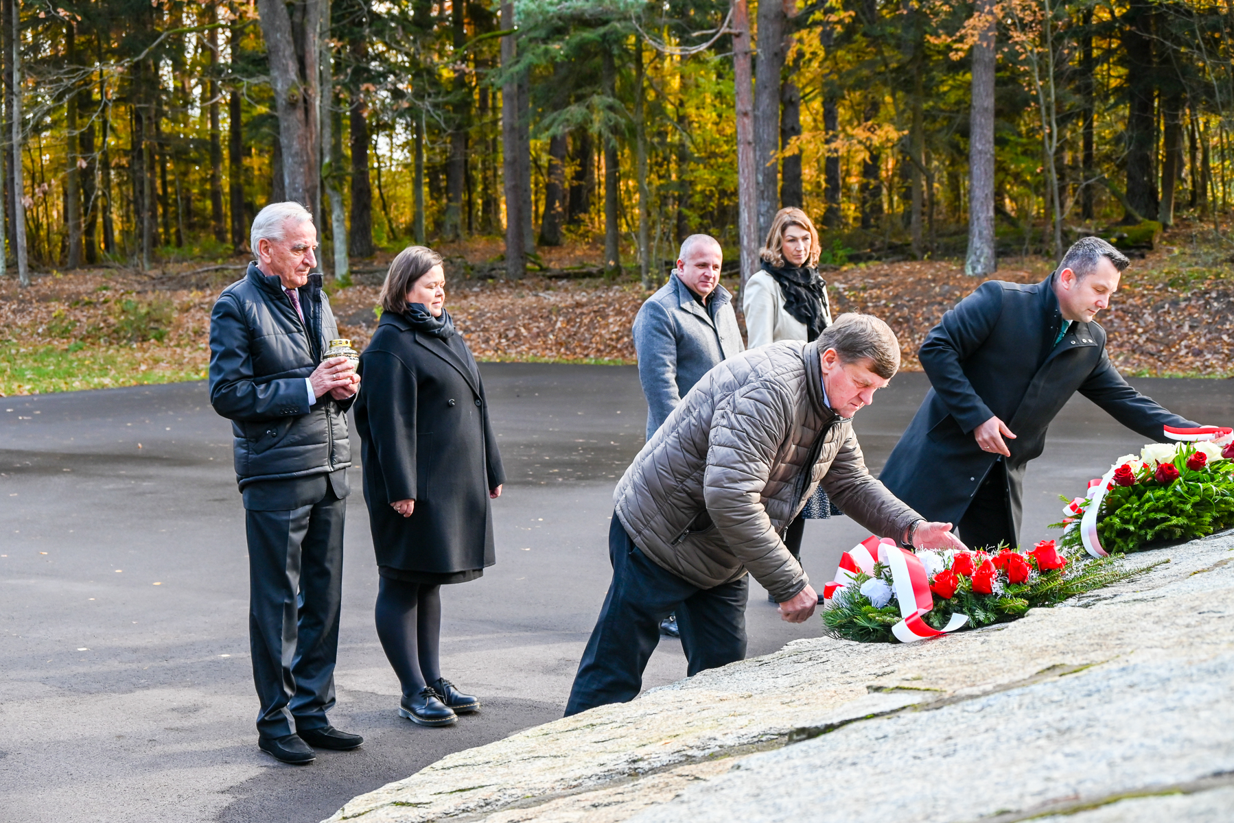 Grupa sześciu osób w jesiennym lesie, dwie składają wiązanki z czerwonych róż i białych kwiatów na kamiennym pomniku, pozostali stoją w zadumie na asfaltowej powierzchni.