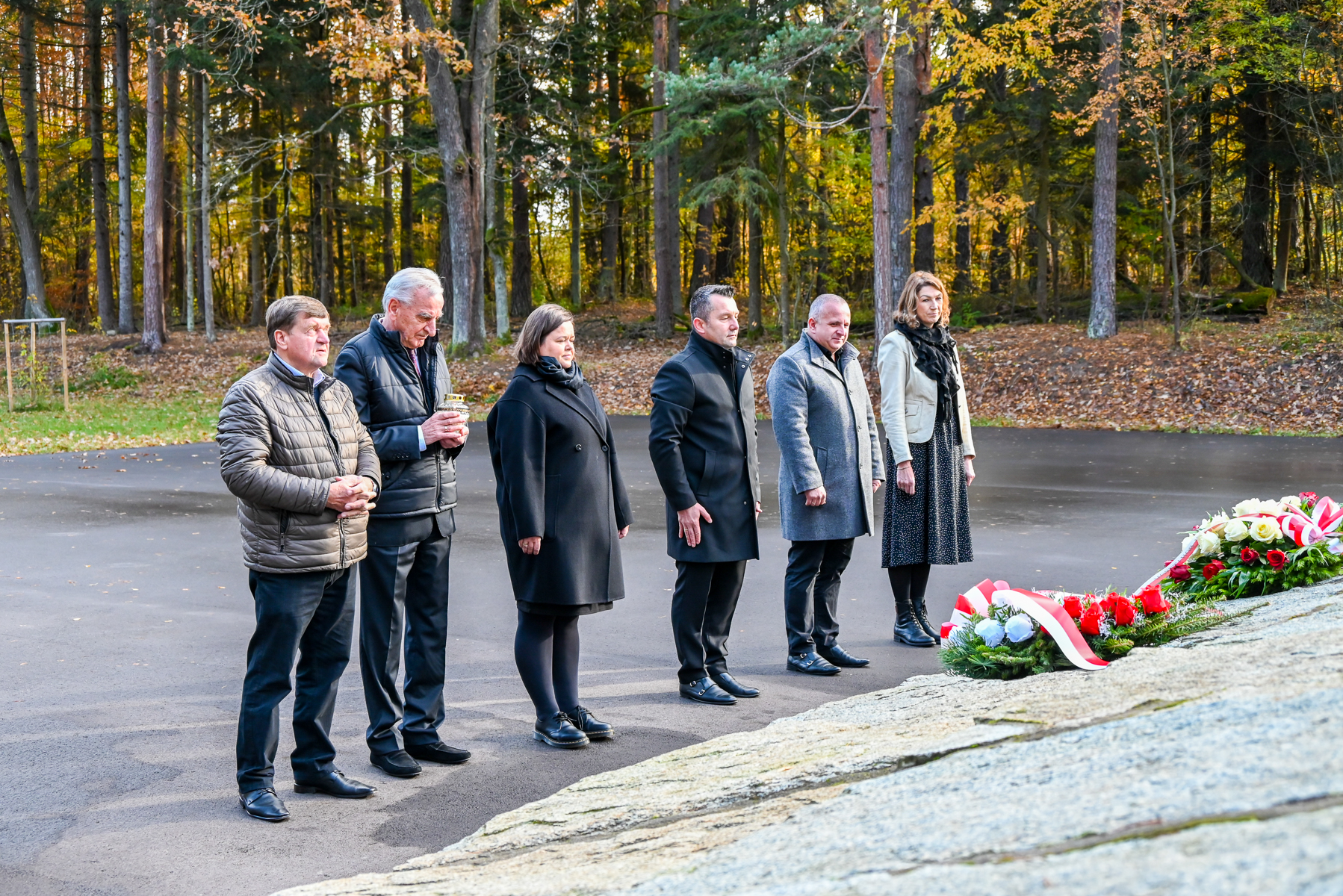 Sześć osób w jesiennym lesie stoi na asfalcie obok kamiennego pomnika z wieńcami z czerwonych i białych kwiatów oraz wstążkami.