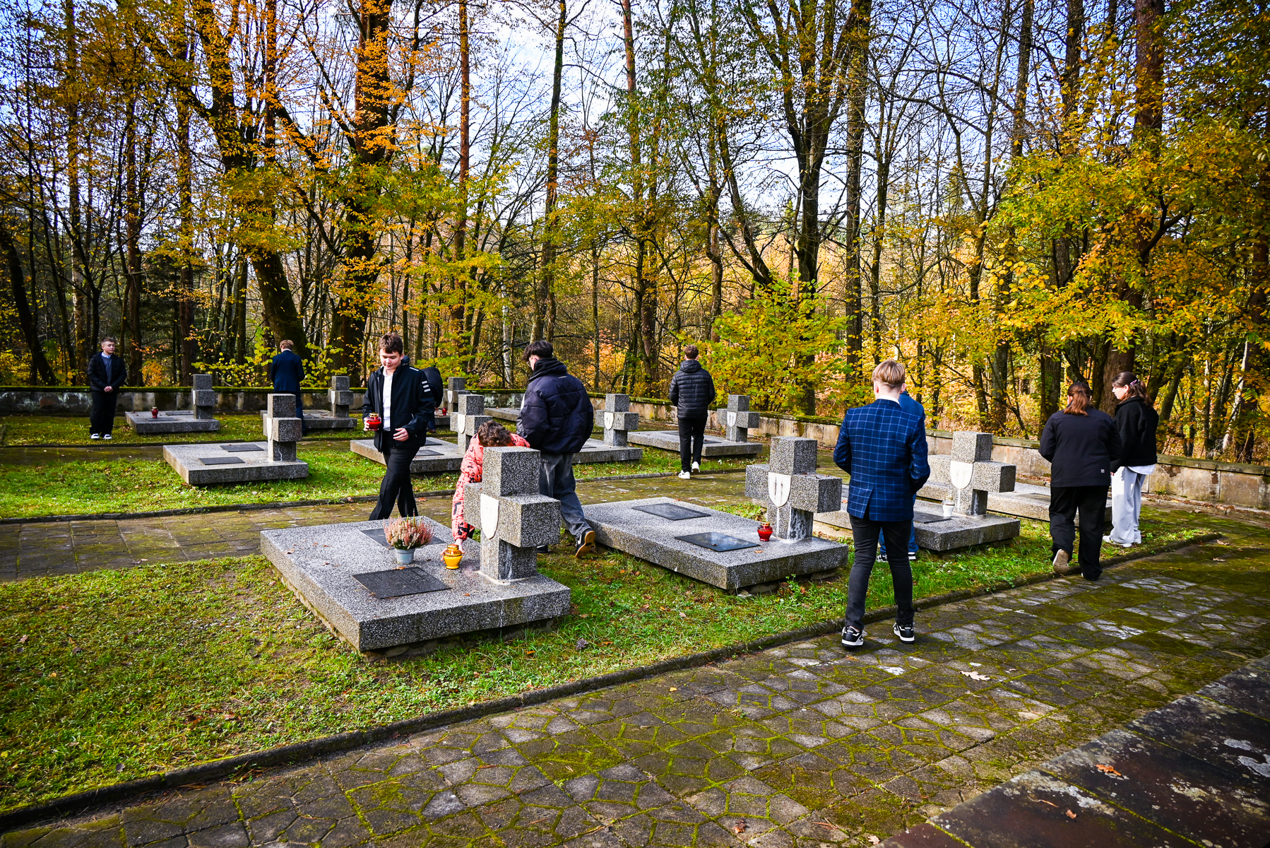 Kilka osób odwiedzających szary cmentarz z granitowymi nagrobkami w lesie z żółto-zielonymi drzewami jesienią.