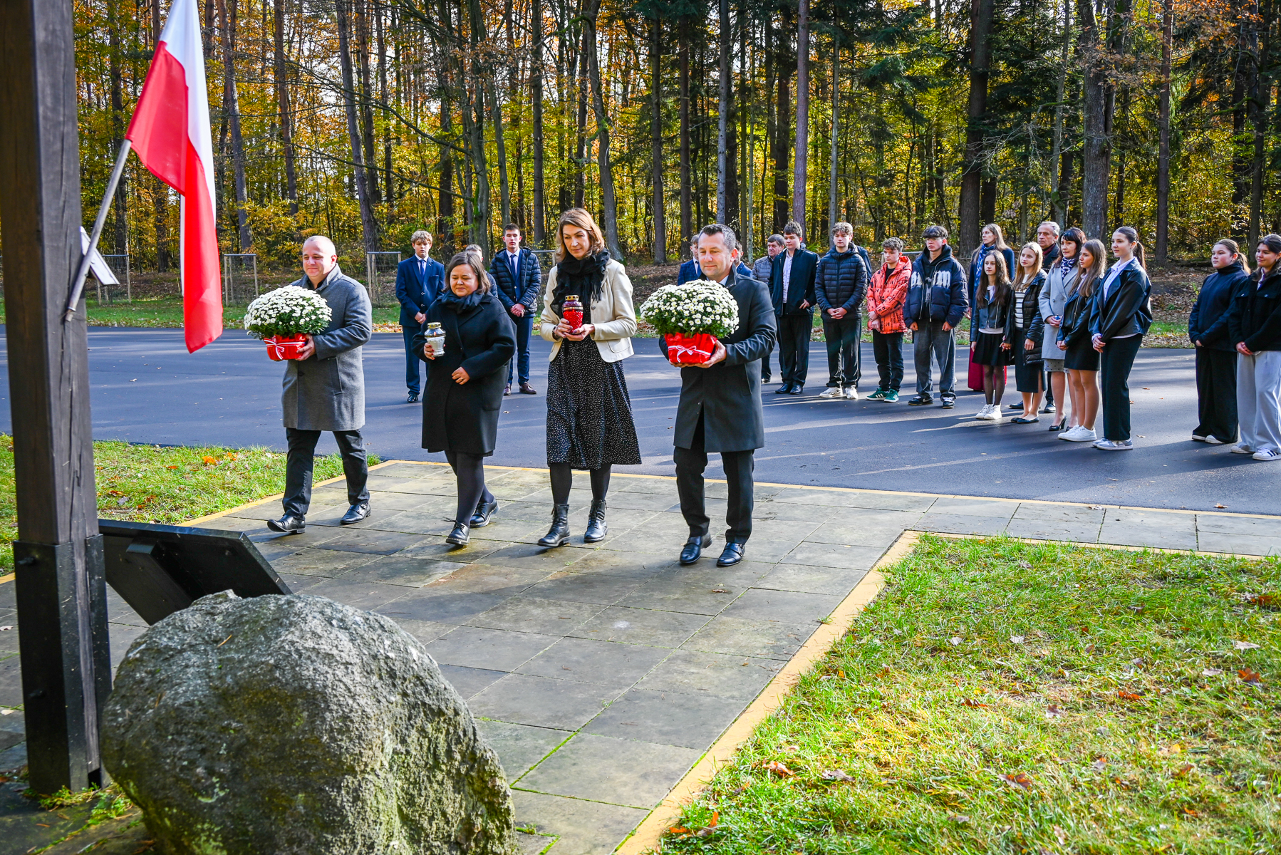 Grupa ludzi w jesiennym lesie, czworo z przodu niesie białe kwiaty i znicze, za nimi stoi kilkanaście osób na asfaltowej nawierzchni, po lewej flaga Polski.
