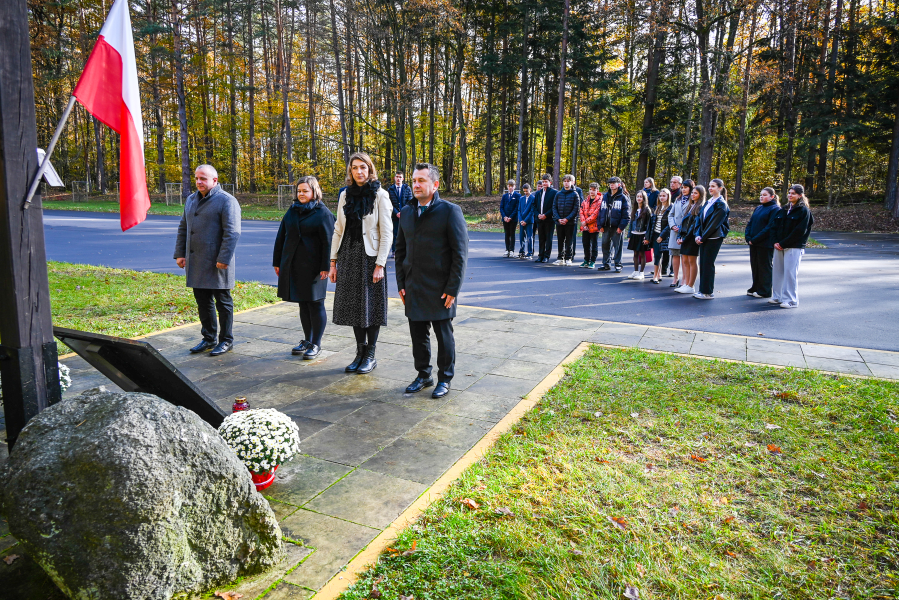 Grupa ludzi stojących na chodniku i asfalcie w lesie, cztery osoby z przodu w ciemnych kurtkach, biało-czerwona flaga, biały kwiat w doniczce obok dużego kamienia.