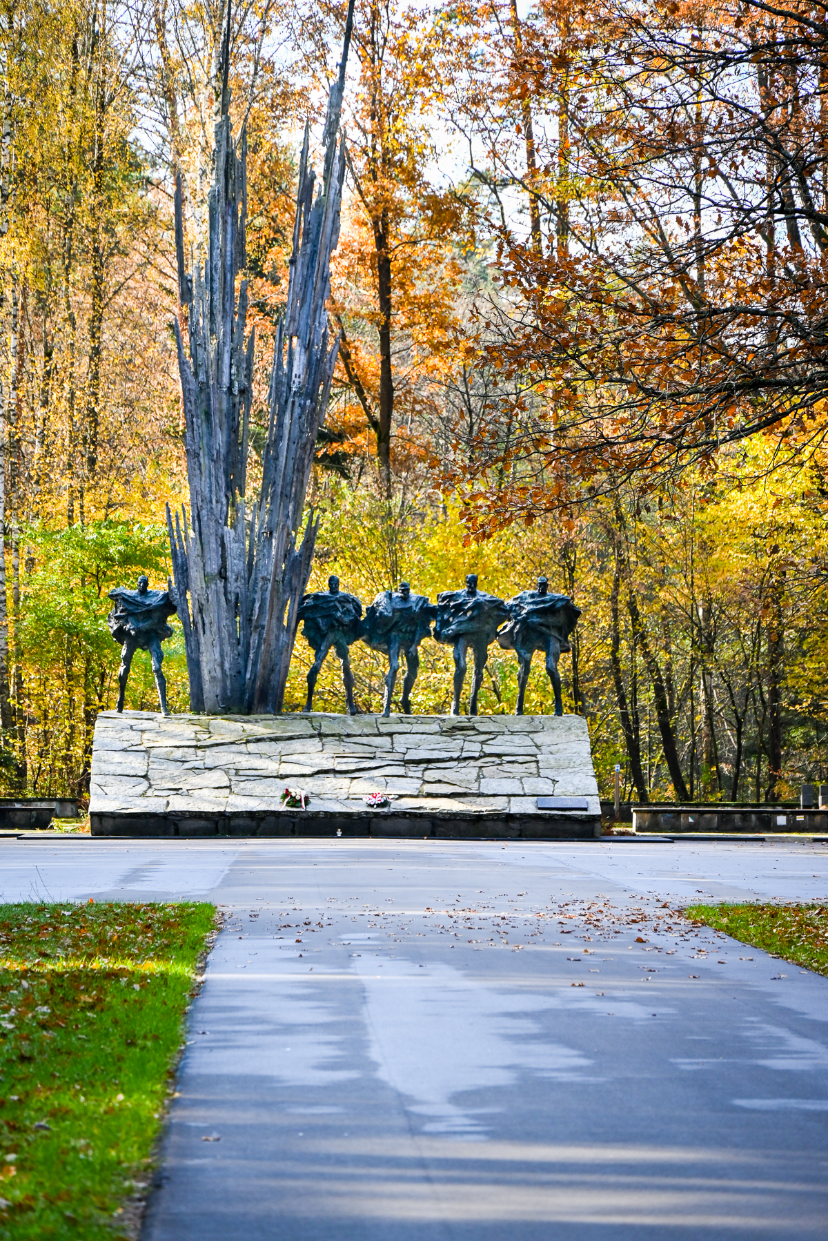 Pomnik z postaciami ludzi i wysokimi, ostrymi formami na kamiennej podstawie, otoczony jesiennym lasem z żółtymi i pomarańczowymi liśćmi, z asfaltową ścieżką na pierwszym planie.