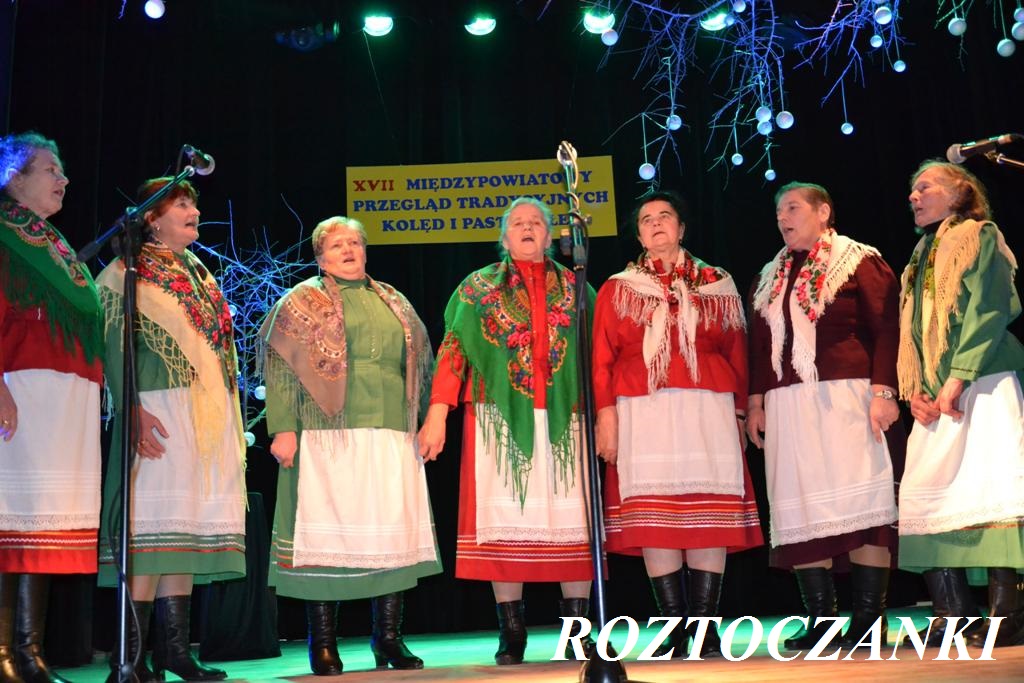 Siedem kobiet w tradycyjnych strojach ludowych z chustami i spódnicami, stojących na scenie pod zielonym oświetleniem, z dekoracjami gałązek i białych kul.