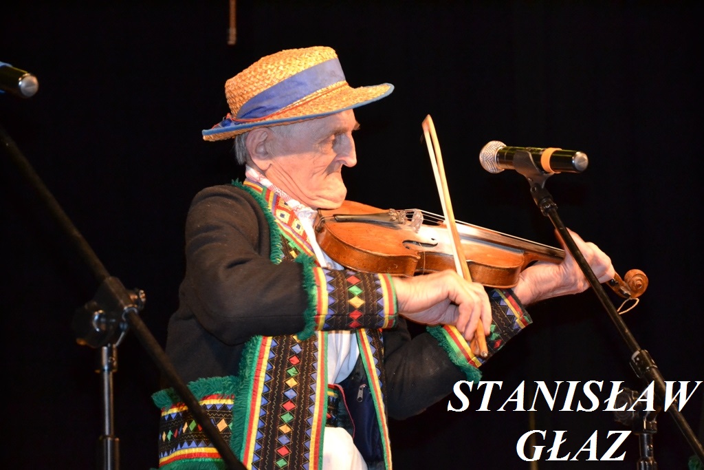 Starszy mężczyzna w czarnym, kolorowym tradycyjnym ubraniu i słomkowym kapeluszu gra na skrzypcach na tle czarnego tła, obok mikrofon na statywie.