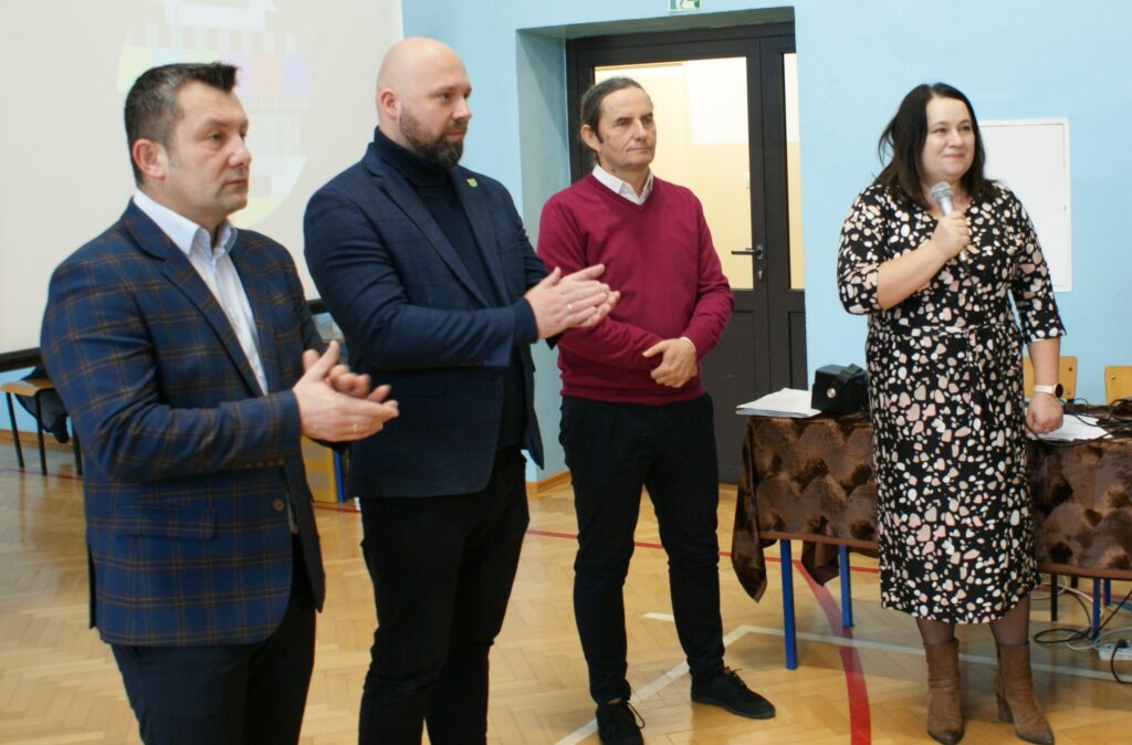 Cztery osoby stoją na sali z jasnobrązową podłogą, trzy mężczyźni w garniturach i swetrze, kobieta w czarno-beżowej sukience trzyma mikrofon.