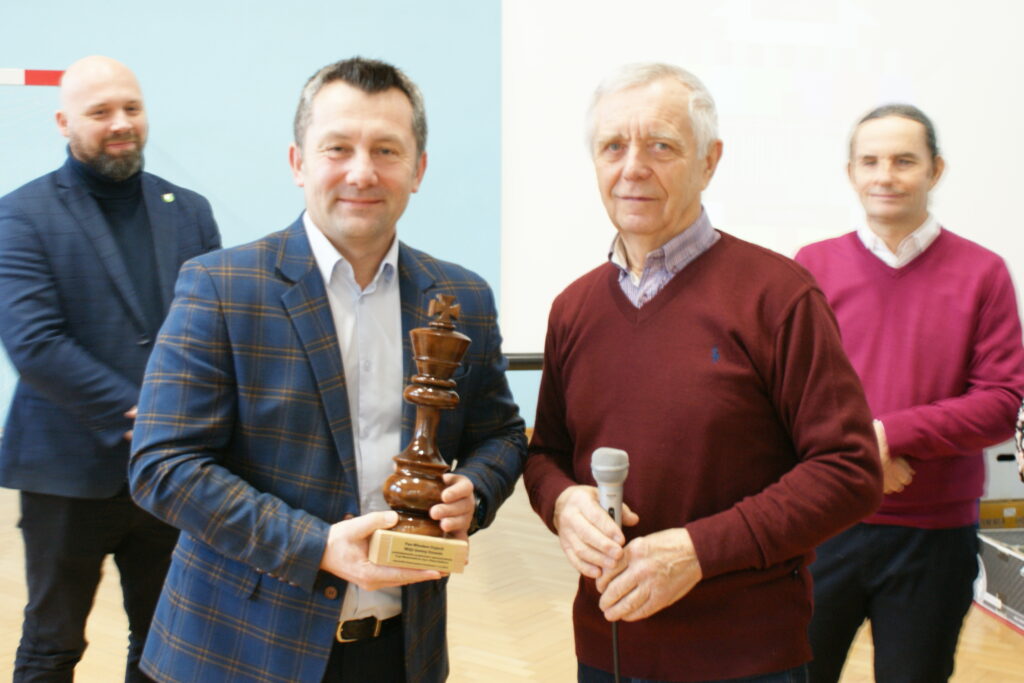 Czterech mężczyzn stojących na sali, dwóch z przodu trzyma mikrofon i dużą drewnianą figurę szachową króla, ubrani w marynarki i swetry w odcieniach granatu, bordowym i różowym.