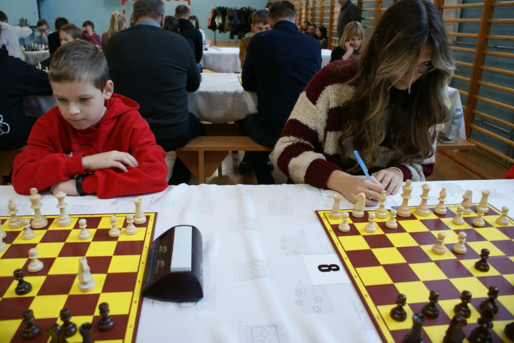 Chłopiec w czerwonej bluzie i dziewczyna w pasiastym swetrze siedzą przy stole z dwoma szachownicami, dziewczyna pisze, w tle ludzie siedzą na ławkach w sali gimnastycznej.