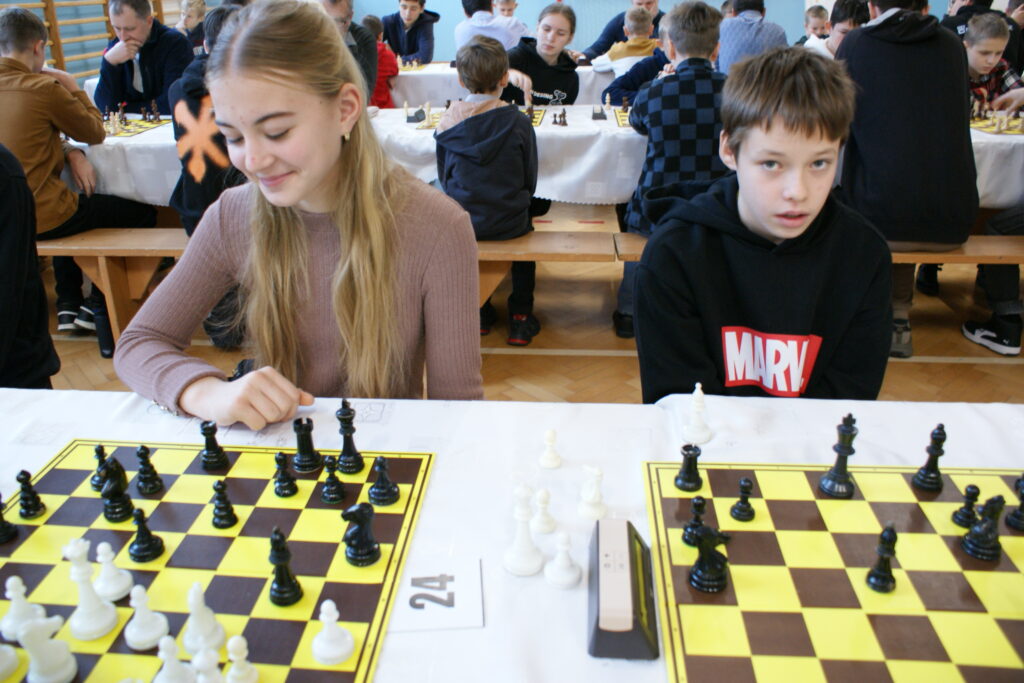 Dwoje dzieci przy stole z szachownicami żółto-brązowymi, dziewczynka w różowym swetrze uśmiechnięta, chłopiec w czarnej bluzie z logo Marvel, w tle więcej grających dzieci.