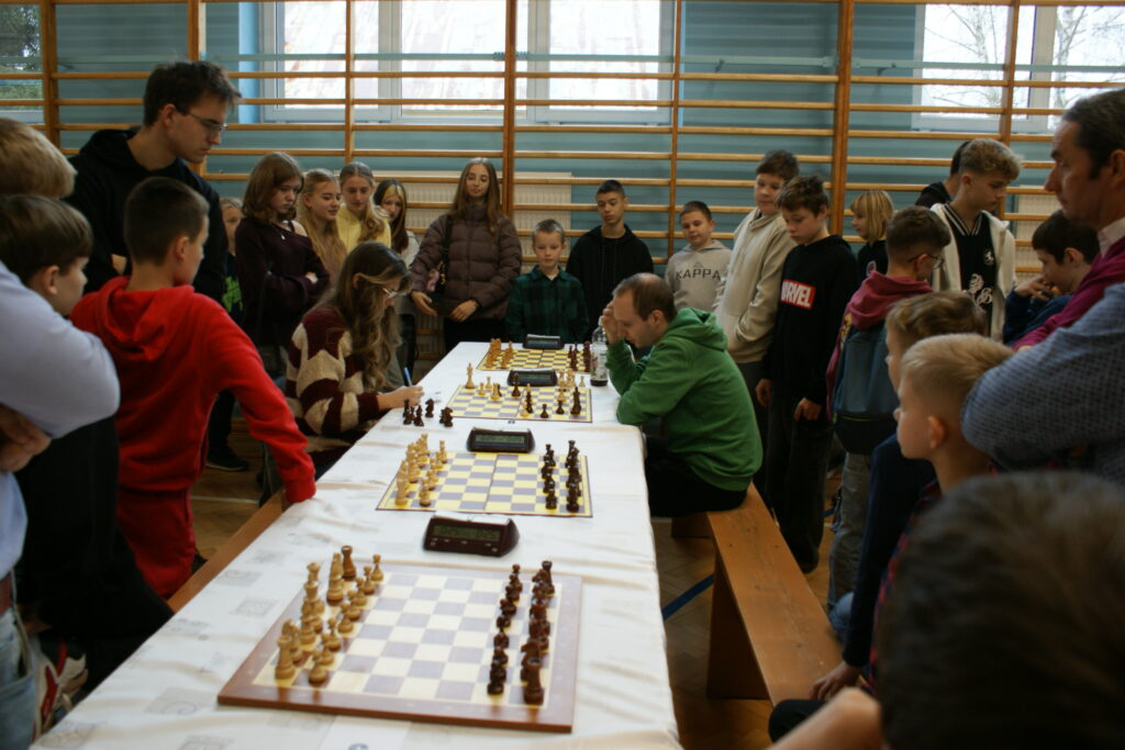 Grupa dzieci i dorosłych wokół stołu z kilkoma szachownicami, dwie osoby siedzą i grają w szachy, wnętrze hali sportowej z drewnianymi drabinkami i oknami.