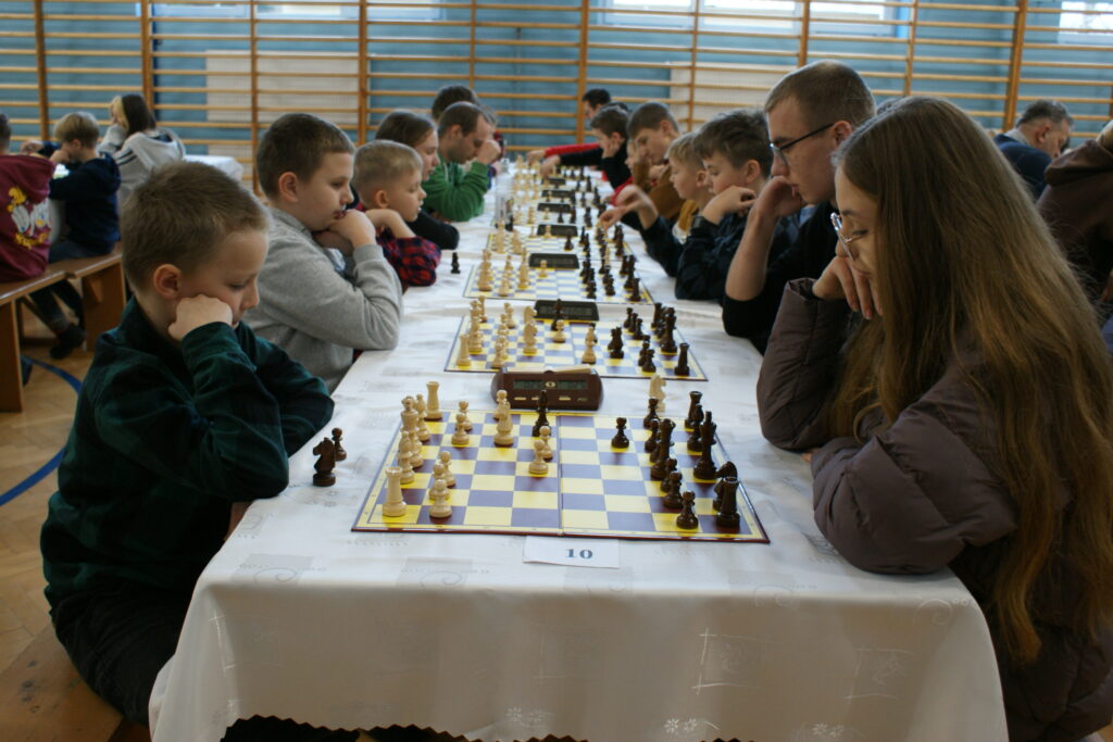 Dzieci i młodzież siedzą naprzeciwko siebie przy długim stole z białym obrusem, grając w szachy na kilku planszach, w tle drewniana ścianka gimnastyczna.