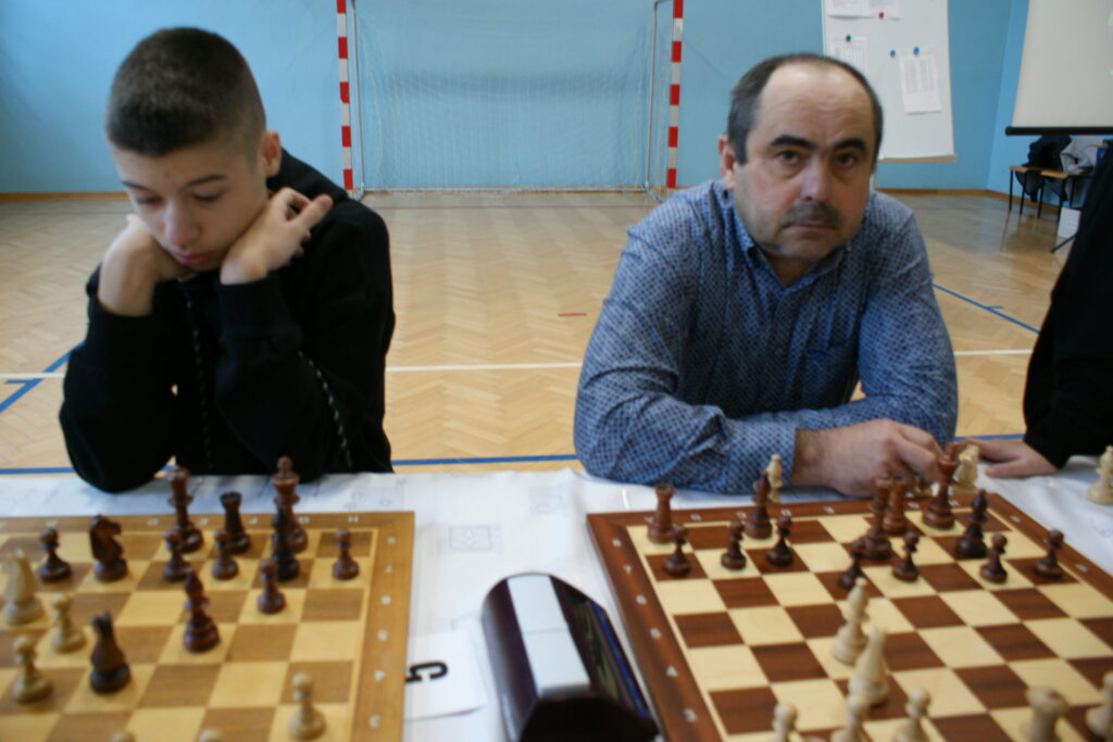 Dwaj mężczyźni siedzą przy stolikach z szachownicami, w tle bramka halowa i jasnoniebieska ściana.