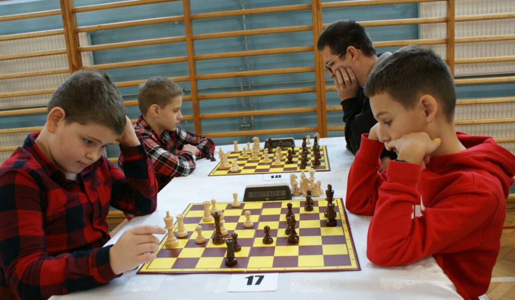 Cztery osoby grające w szachy przy dwóch planszach na białym stole, dwie osoby po lewej w czerwono-czarnych ubraniach, dwie po prawej, jedna w czarnym, druga w czerwonym.