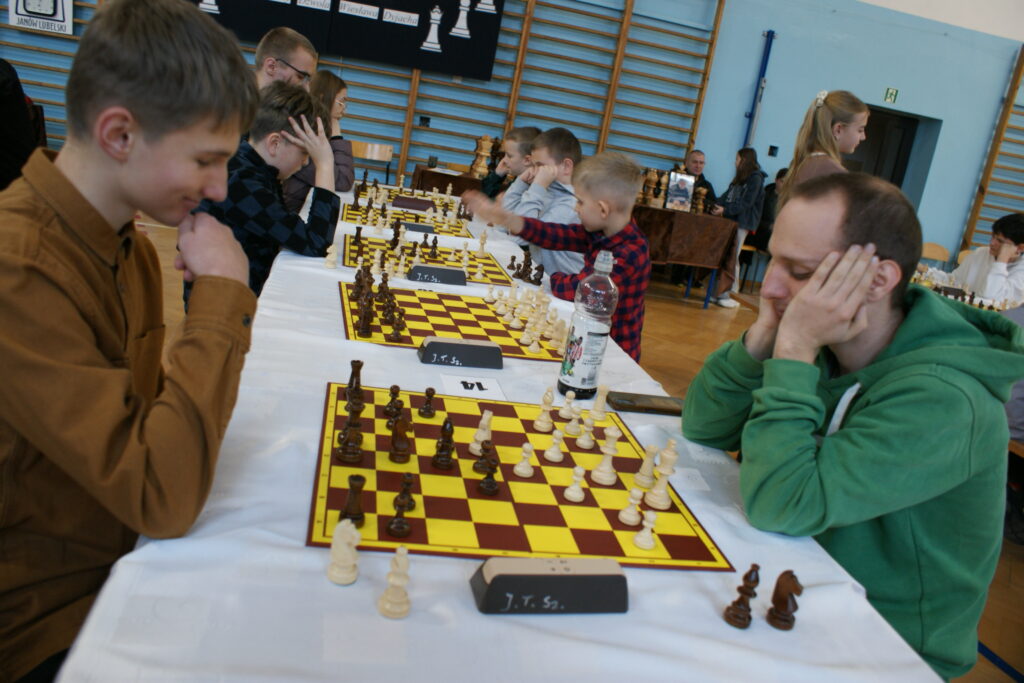 Kilka osób różnego wieku grających w szachy przy długim stole z białym obrusem w jasnoniebieskiej sali gimnastycznej.