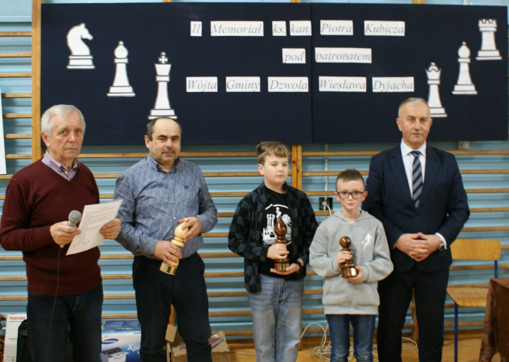 Pięć osób w hali sportowej na tle planszy z dużymi szachowymi figurami i napisem, dwie osoby dorosłe trzymają mikrofon i kartkę, dwie dzieci trzymają drewniane trofea.