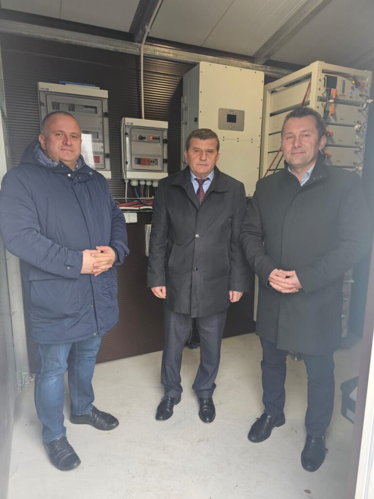 Trzech mężczyzn w ciemnych płaszczach stoi na jasnej podłodze przed ścianą z szarymi i białymi urządzeniami elektrycznymi i kablami.