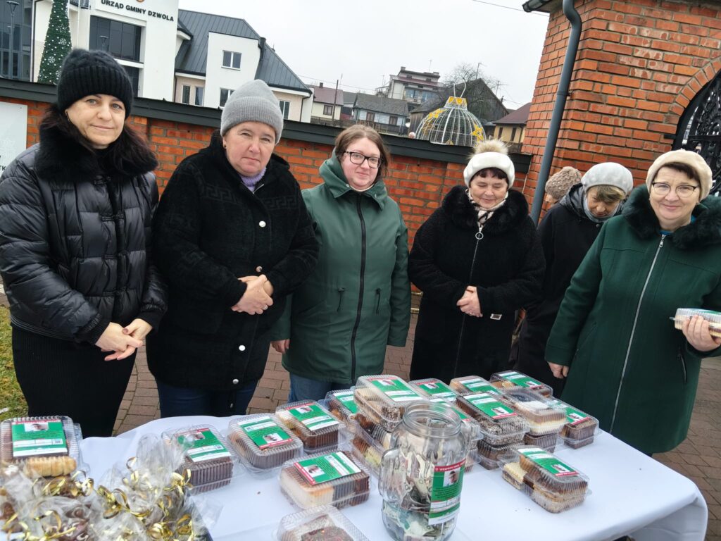 Sześć kobiet w zimowych kurtkach i czapkach stoi za stołem z plastikowymi pojemnikami z ciastami i przezroczystym słoikiem z pieniędzmi, na tle ceglanej ściany i budynków.