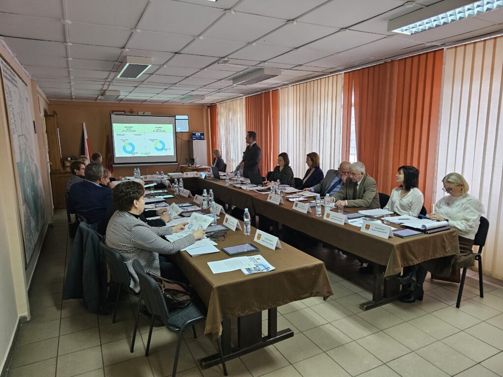 Ludzie siedzący przy dwóch stołach z brązowymi obrusami, na ścianie ekran z wykresami i okna z pionowymi zasłonami w odcieniach pomarańczowego i beżu.