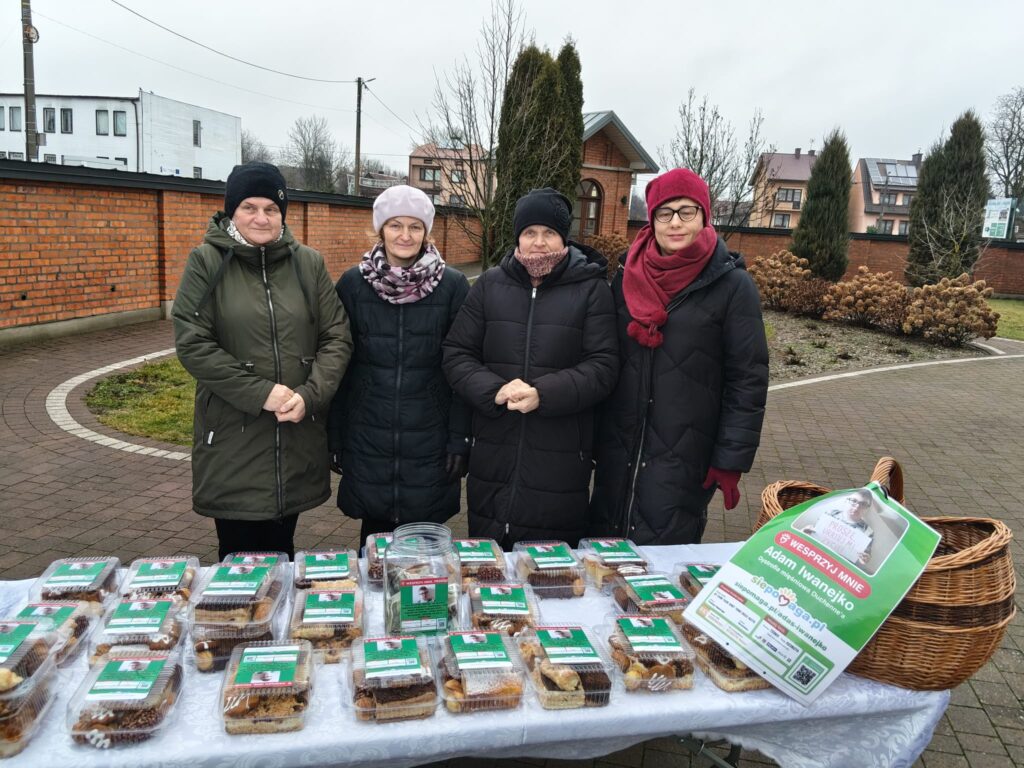 Cztery kobiety w zimowych kurtkach i czapkach stoją za stołem z opakowaniami ciast na zewnątrz, obok kosza wiklinowego i plakatu z prośbą o wsparcie.