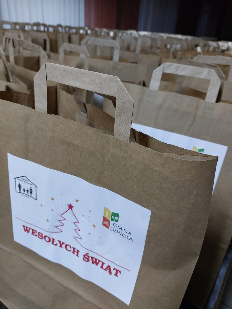 Brązowe papierowe torby z uchwytami, na których znajdują się białe naklejki z napisem "WESOŁYCH ŚWIĄT" i symbolami. Torby ułożone w rzędach.