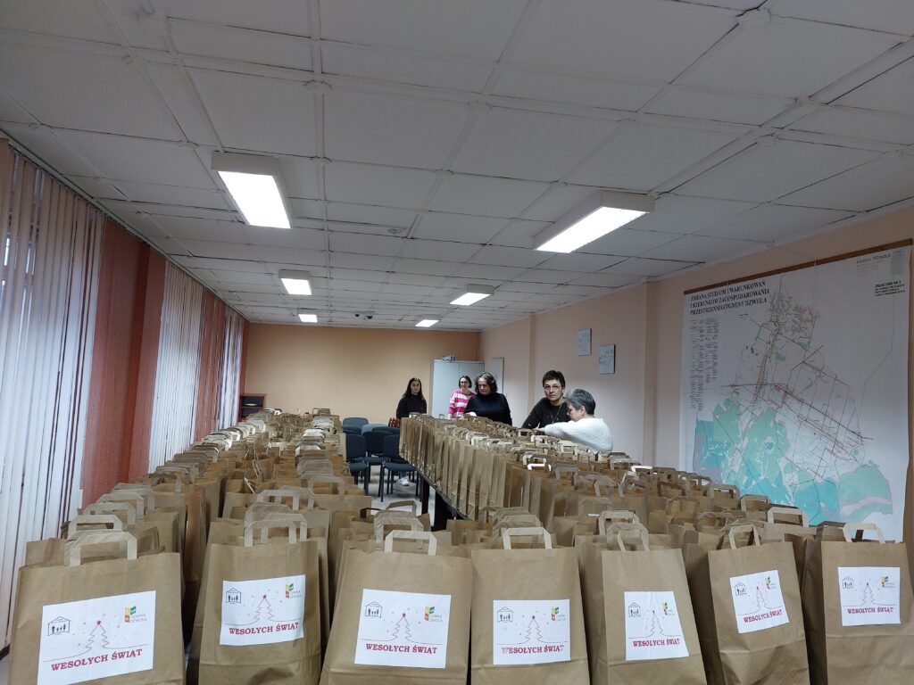 Sześć kobiet stojących przy stolikach z dużą ilością brązowych papierowych toreb z napisem "Wesołych Świąt" w jasnym pomieszczeniu z mapą na ścianie.