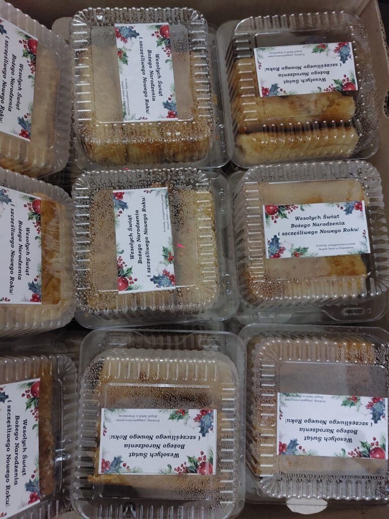 Przezroczyste plastikowe pojemniki z prostokątnymi ciastami, na których znajdują się białe naklejki z tekstem i motywem świątecznym.