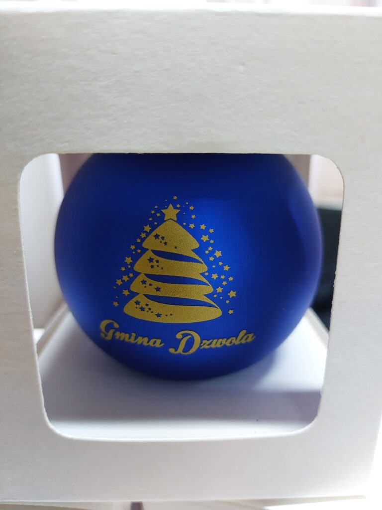 Niebieska matowa bombka z nadrukiem złotego choinkowego wzoru i napisu "Gmina Dzwola" w białym pudełku z okienkiem.