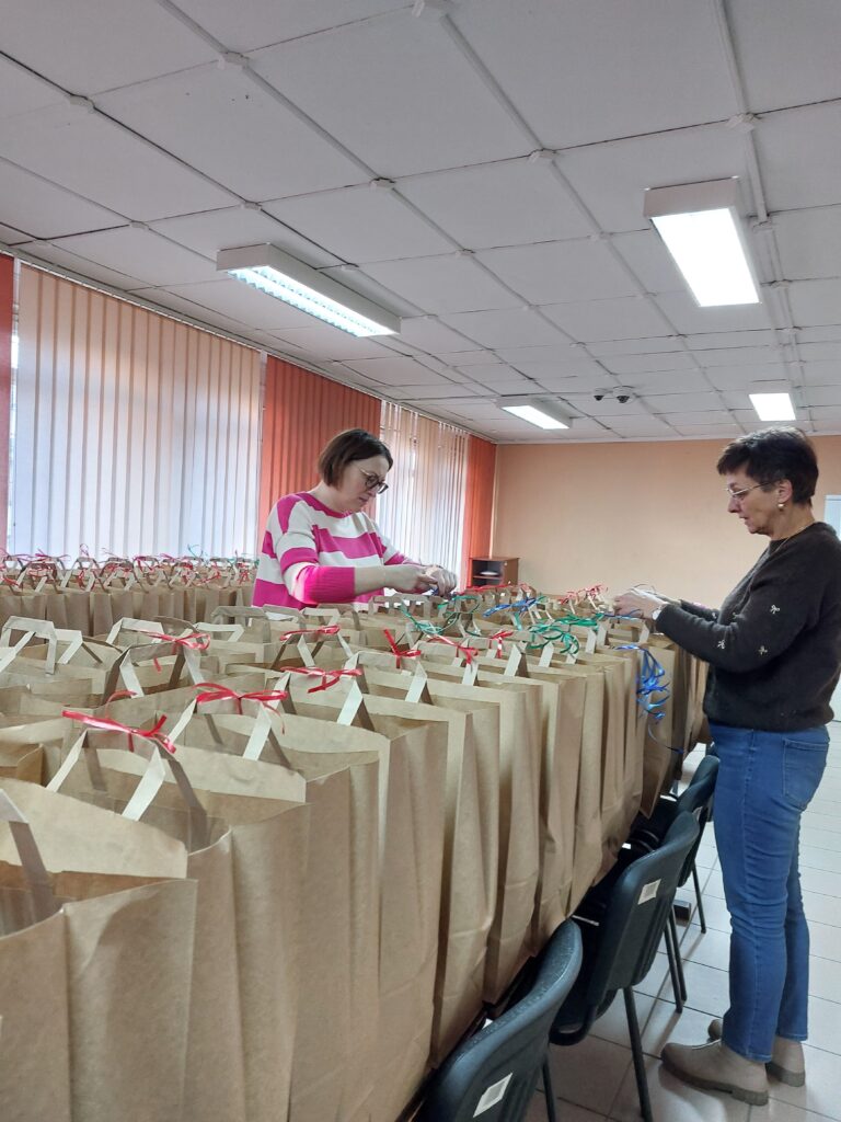 Dwie kobiety przy stole wypełnionym papierowymi torbami z uchwytami, każda torba ozdobiona kolorową wstążką, wnętrze z jasnym sufitem i pomarańczowymi zasłonami.