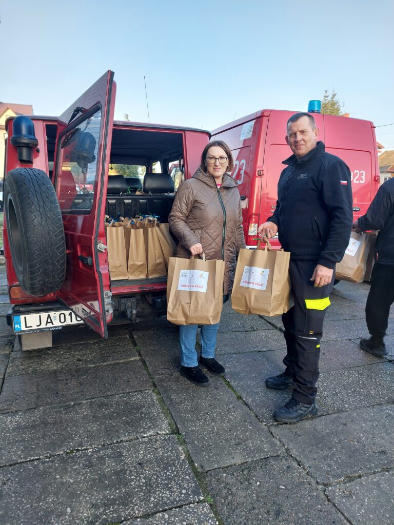 Kobieta i mężczyzna stojący przy otwartym czerwonym samochodzie, oboje trzymają papierowe torby z napisem "Wesołych Świąt". W tle drugi czerwony pojazd.