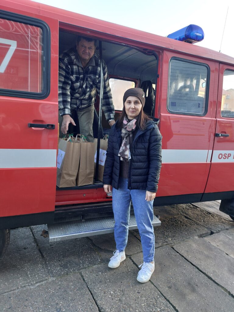 Człowiek w czarnej kurtce, niebieskich jeansach i brązowej czapce stoi obok czerwonego pojazdu z napisem OSP, w środku mężczyzna w koszuli w kratę trzyma papierowe torby.