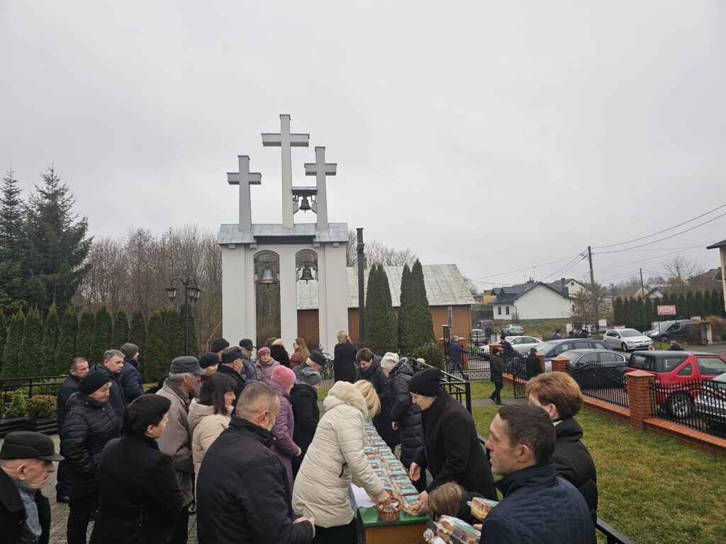 Grupa ludzi w zimowych ubraniach stojąca przy stołach z paczkami jedzenia na zewnątrz, w tle biała dzwonnica z trzema krzyżami i dzwonami, pochmurne niebo.