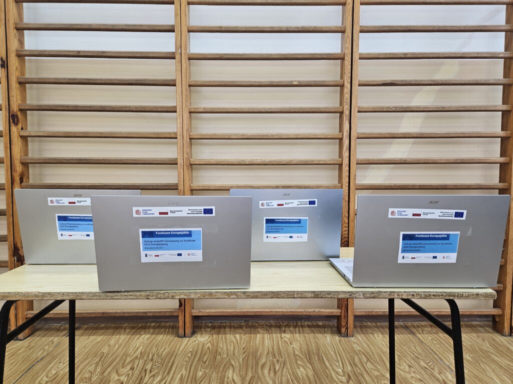 Cztery srebrne laptopy Acer z naklejkami informacyjnymi, ustawione na jasnym stole, w tle drewniane drabinki gimnastyczne i jasna ściana.