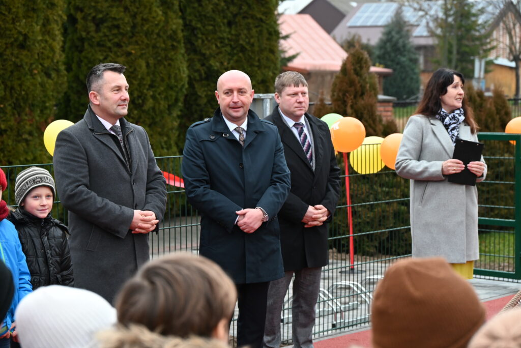 Czterech dorosłych w płaszczach i dzieci na zewnątrz, za nimi zielone drzewka i kolorowe balony przy metalowym ogrodzeniu.