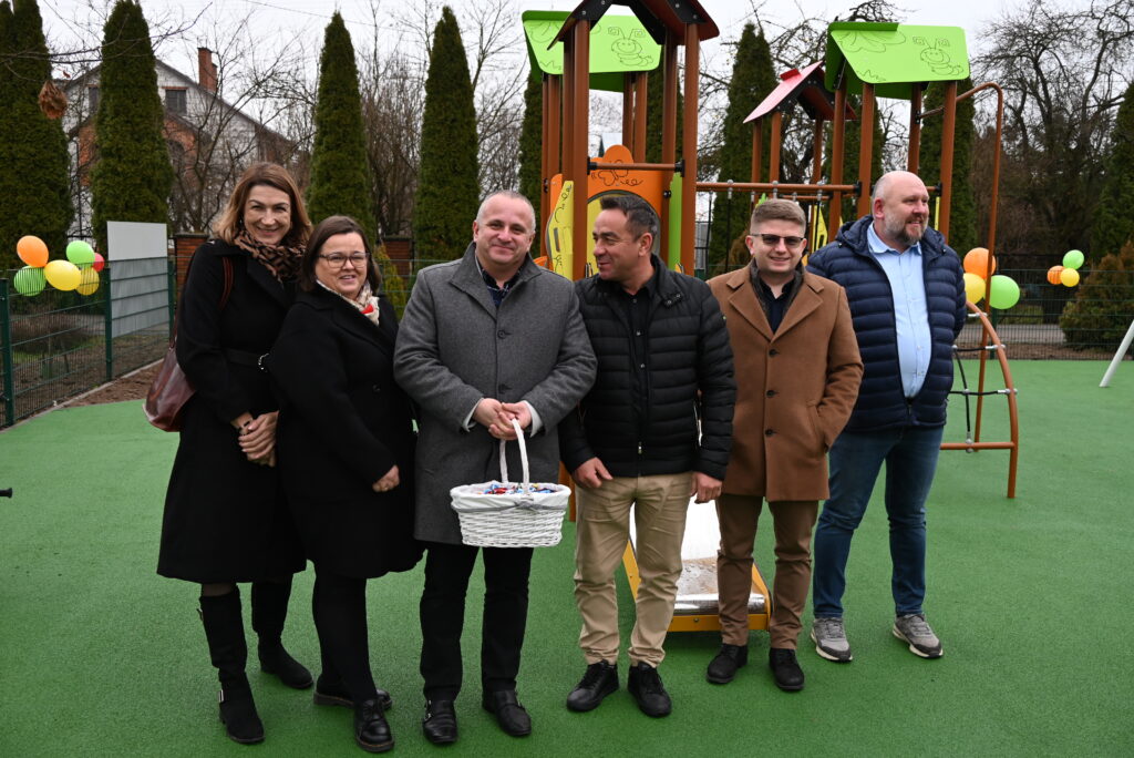 Grupa sześciu osób stoi na zielonej nawierzchni placu zabaw, w tle kolorowa konstrukcja do zabawy i balony przy ogrodzeniu.