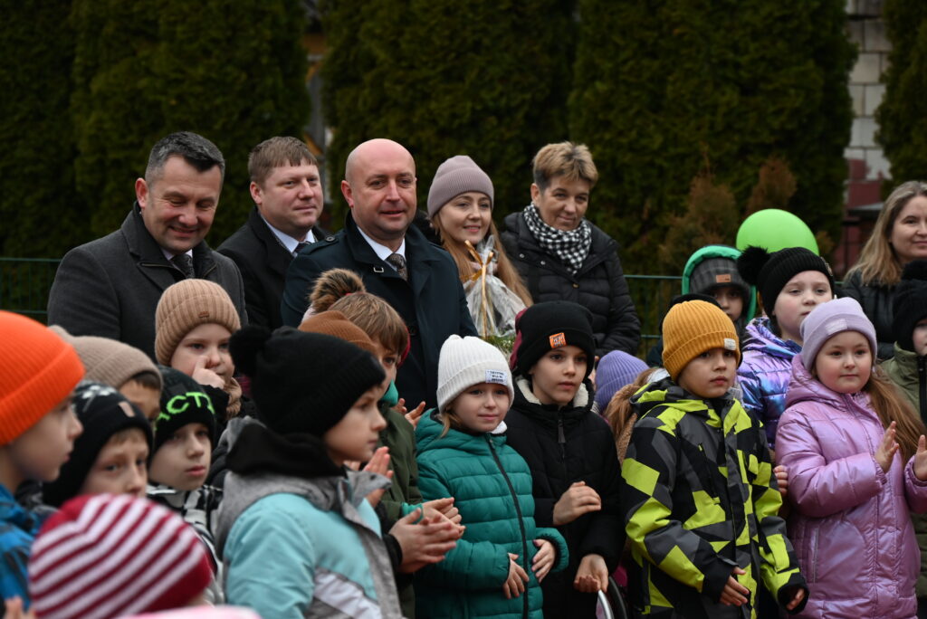 Grupa dzieci i dorosłych ubranych w zimowe kurtki i czapki stoi na tle zielonych drzew. Niektóre dzieci klaszczą, jedna osoba trzyma prezent w przezroczystym opakowaniu.