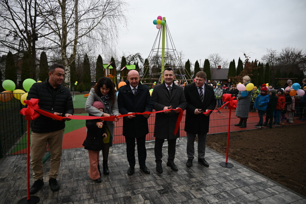 Pięć osób tnie czerwoną wstęgę na tle placu zabaw z zielono-pomarańczową nawierzchnią, balonami i płotem, w oddali grupa dzieci i dorosłych.