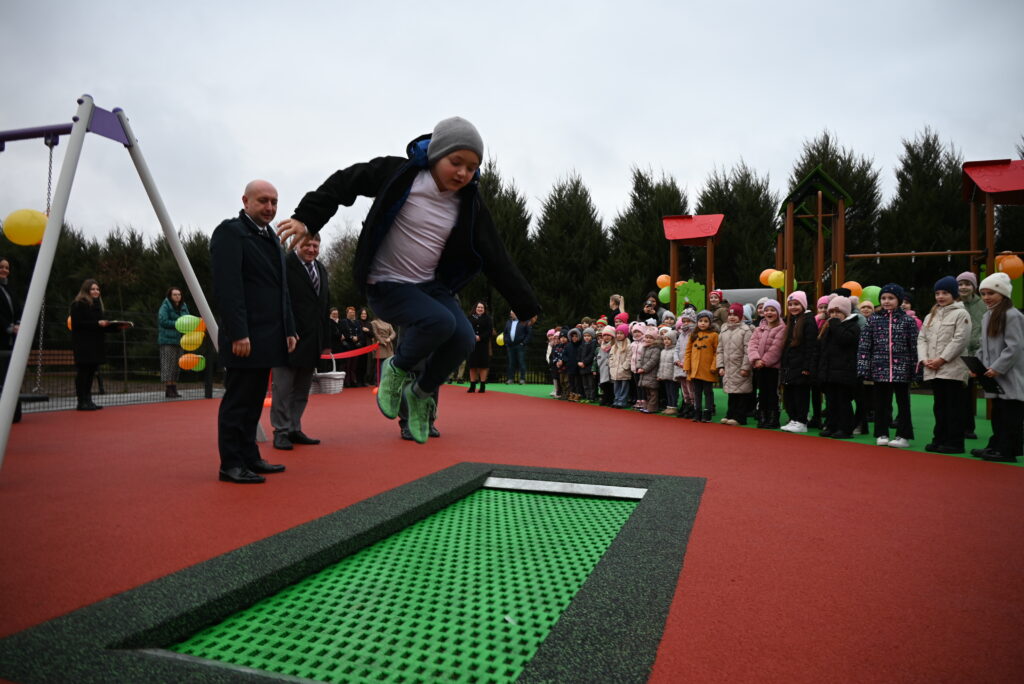 Chłopiec w czapce i zielonych butach skacze na zielonej trampolinie w otoczeniu dzieci i dorosłych na placu zabaw z czerwonym i zielonym podłożem, w tle drzewa i zabawki.