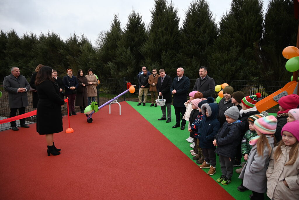 Grupa dorosłych i dzieci stojących na placu zabaw z czerwoną i zieloną nawierzchnią, z huśtawką wahadłową i zjeżdżalnią, w tle wysokie zielone drzewa.