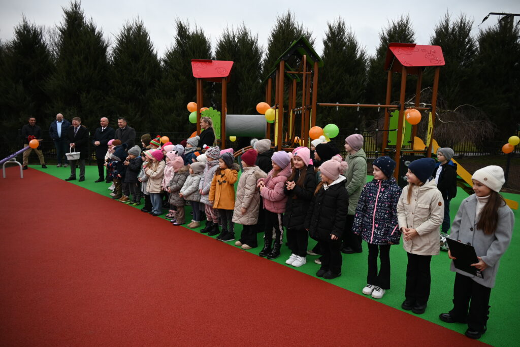 Grupa dzieci i kilku dorosłych stoi na placu zabaw z zieloną i czerwoną nawierzchnią, w tle drewniana konstrukcja zabawowa z czerwonymi daszkami i kolorowymi balonami.