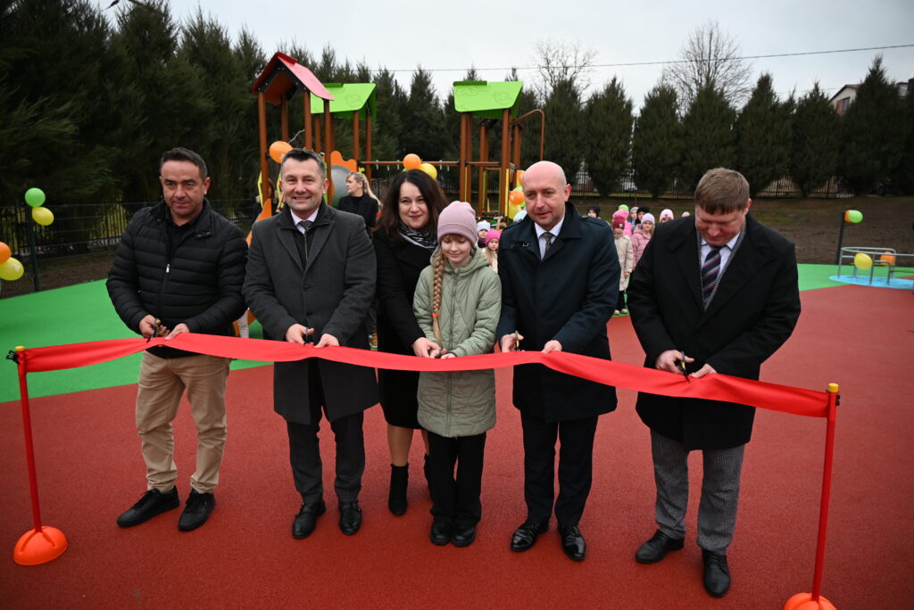 Pięć osób, w tym dziewczynka w zielonej kurtce i różowej czapce, przecina czerwoną wstęgę na tle placu zabaw z zielono-pomarańczową konstrukcją i grupą dzieci w pastelowych ubraniach.
