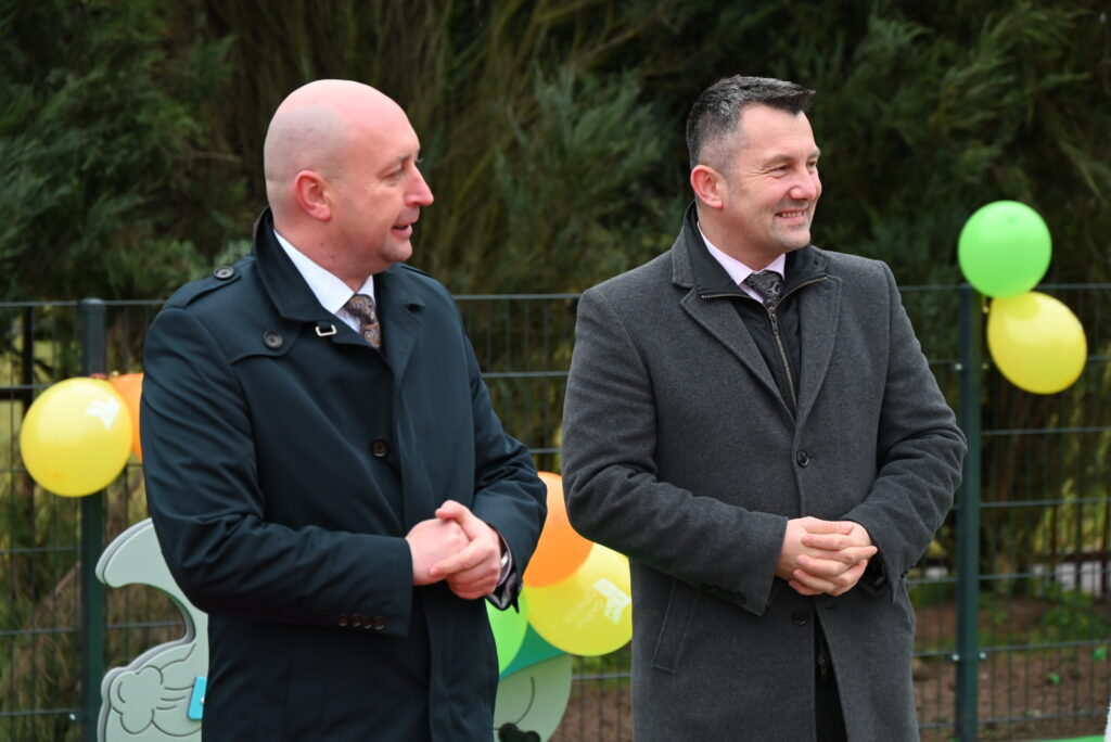 Dwaj mężczyźni w ciemnych płaszczach i krawatach stoją na tle ogrodzenia z zielonymi i żółtymi balonami.