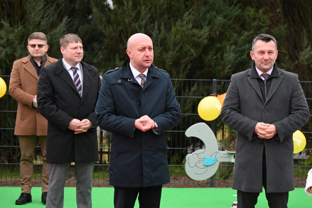 Czterech mężczyzn w płaszczach stoi na zielonym placu zabaw, w tle ogrodzenie, balony i element zabawki w kształcie ptaka.