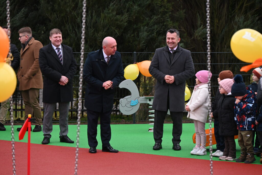 Grupa mężczyzn w ciemnych płaszczach i dzieci w zimowych ubraniach stoją na placu zabaw z zieloną i czerwoną nawierzchnią, ozdobionym żółtymi i pomarańczowymi balonami.