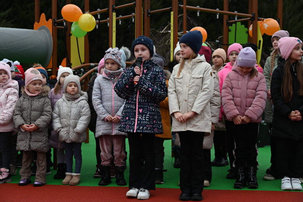 Grupa dzieci w zimowych ubraniach stoi na placu zabaw z zieloną i czerwoną nawierzchnią, jedna dziewczynka trzyma mikrofon, w tle kolorowe balony i elementy placu zabaw.