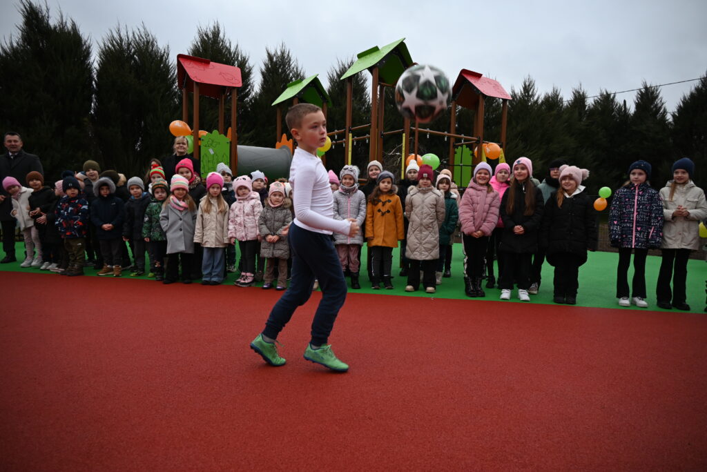 Chłopiec w białej bluzie i zielonych butach kopie piłkę na czerwonym placu, w tle grupa dzieci w kolorowych kurtkach i czapkach oraz plac zabaw z zielonym i czerwonym daszkiem.